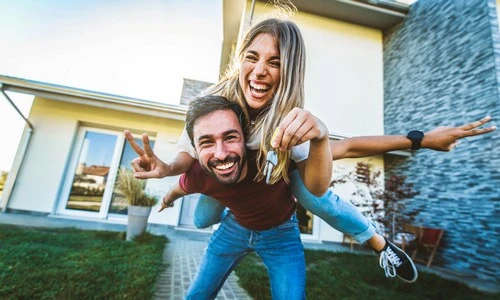 Hypothek Waadt: Glückliches Paar (Mann trägt seine Frau auf dem Rücken) vor ihrem mit einem Hypothekarkredit gekauften Haus